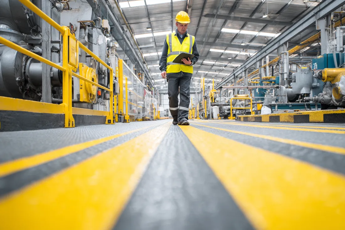 A health-and-safety manager in hi-vis and hard hat walking a marked anti-slip safety walkway in a workshop